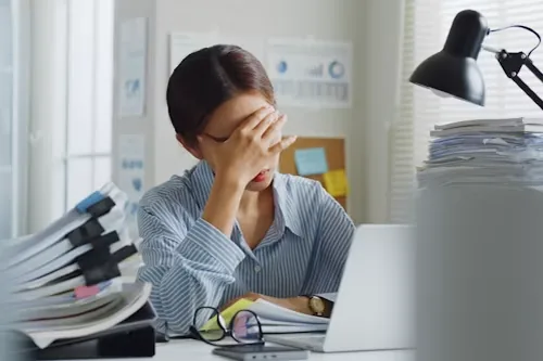 A person at a desk with stacks of paperwork, head in hand