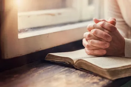 Clasped hands resting on an open book beside a sunlit window