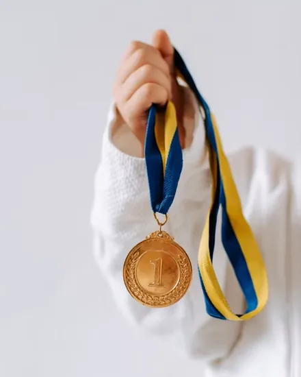A hand holding out a gold first-place medal on a blue and yellow ribbon